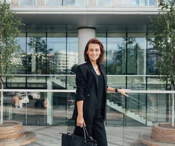 Smiling businesswoman in black formal wear holding a bag standing against an office building  Middle aged female standing outdoors