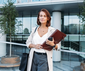 Middle aged female with folder wearing stylish clothes standing outdoors against office building
