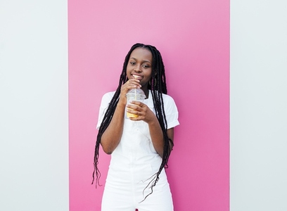Young woman with braids holding a cocktail standing at a white wall with a pink stripe