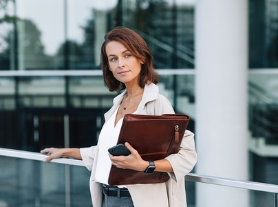 Confident middle aged woman in stylish clothes standing outdoors  Female with ginger hair holding a leather folder while standing against an office building