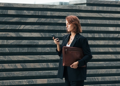 Side view of middle aged businesswoman with leather folder and smartphone walking outdoors