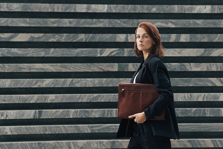 Side view of a confident businesswoman with a leather folder walking outdoors  Middle aged businesswoman in black formal clothes looking away while walking