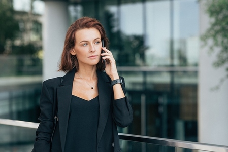 Confident businesswoman talking on mobile phone and looking away  Middle aged female in black formal clothes making conversation on smartphone against glass building
