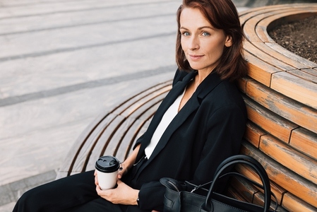 Middle aged smiling businesswoman sitting on a bench outdoors  Female in black formal wear relaxing outdoors holding a coffee cup