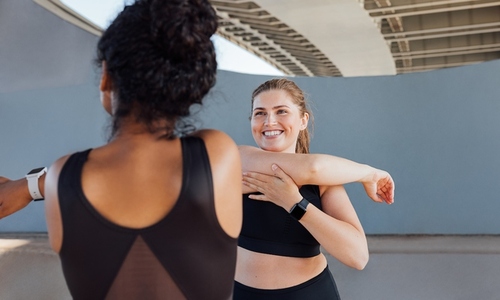 Plus size female smiling and looking at her fitness buddy while stretching her arm  Two women in black fitness attire warming up hands before a workout