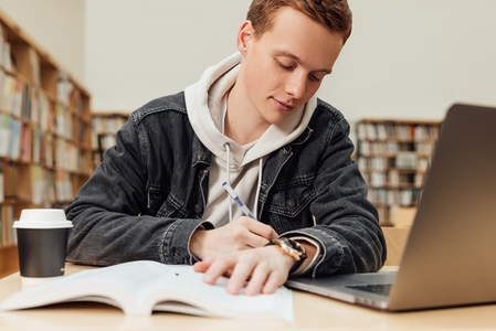 Male student in library writing on notebook while sitting at a table in the library