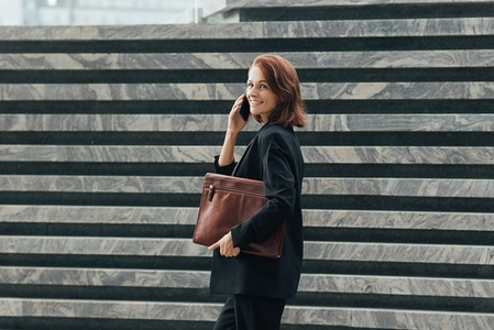 Happy middle aged businesswoman talking on a smartphone while walking  Smiling female in black formal clothes looking back while talking on a smartphone