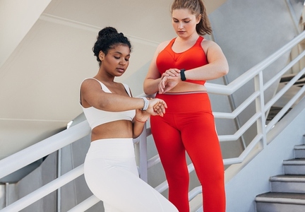 Two plus size women in fitness attire are checking pulse during a workout  Young females in sports clothes are standing on stairs outdoors looking at smartwatches