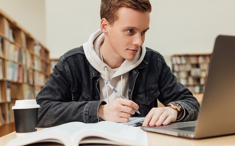 Male student with ginger hair preparing for an exam while sitting in a library  High school student sitting at a table with laptop