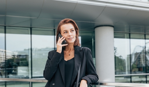 Middle aged female with ginger hair looking away while talking on her mobile phone  Confident woman in formal wear standing at the office building