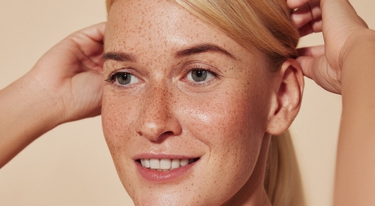 Close up highly detailed portrait of a young smiling woman with freckles  Cropped image of a beautiful blond female adjusting hair against a beige backdrop