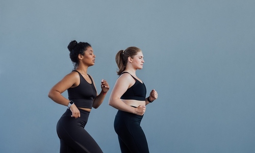 Side view of two females with similar body types running together at grey wall  Two women in black fitness attire running outdoors