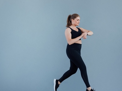 Side view of young plus size female checking smart watch while jogging at a grey wall  Woman in black sportswear looking at her smartwatch while running