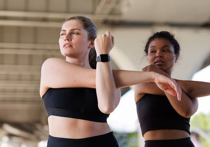 Two females stretch their hands while practicing under a bridge  Young plus size woman doing warming up exercises with her friend and looking away