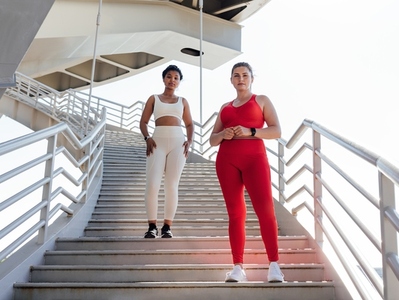 Two females in fitness attire with different colors looking at camera while standing on a staircase  Plus sized woman with her fitness friend posing together