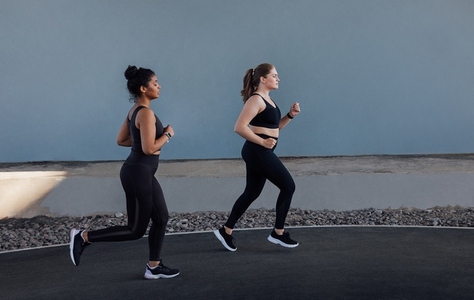 Two women with different body types running outdoors  Side view of two young females jogging at a grey wall