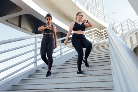 Two young females go on the stairs and watch on their smartwatches  Plus sized woman with her friend looking at smartwatch while stepping down on stairs