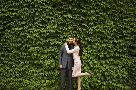 Woman Kissing Man on Cheek in front of Green Leafy Hedge
