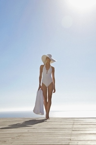Woman in white hat and swimsuit standing on a sun deck holding a towel