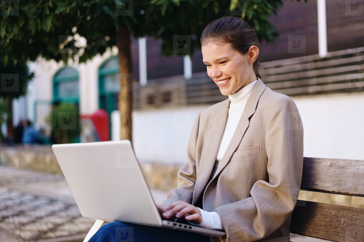 A Young Woman Happily Working on Her Laptop Outdoors While Sitting on a Bench in the Park