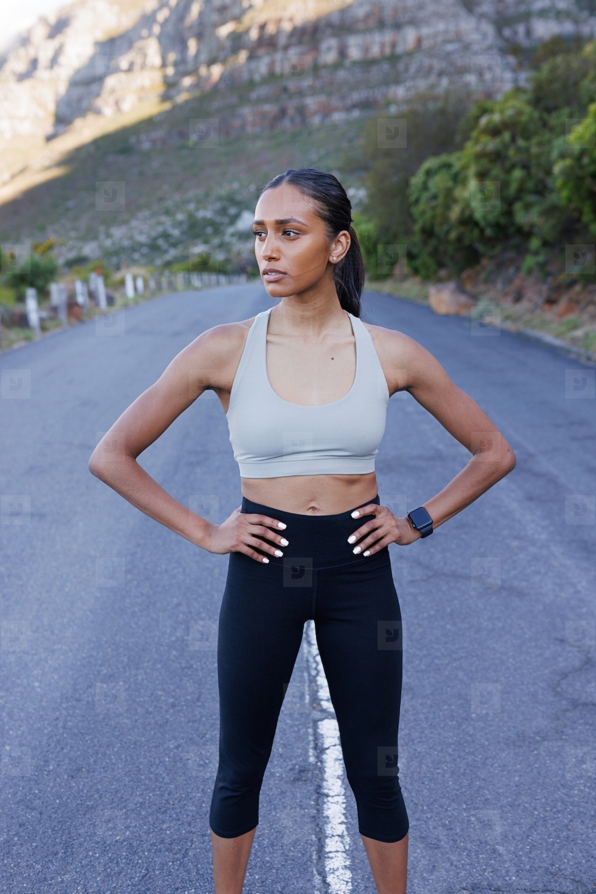 Young confident slim female with hands on her hips standing outdoors on an abandoned road and looking away  Woman jogger relaxing during outdoor workout