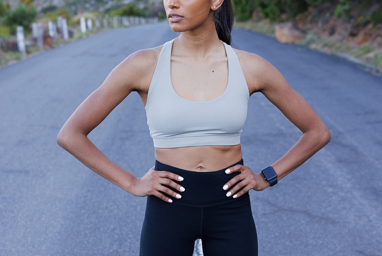 Young confident slim female with hands on her hips standing outdoors on an abandoned road and looking away. Woman jogger relaxing during outdoor workout.