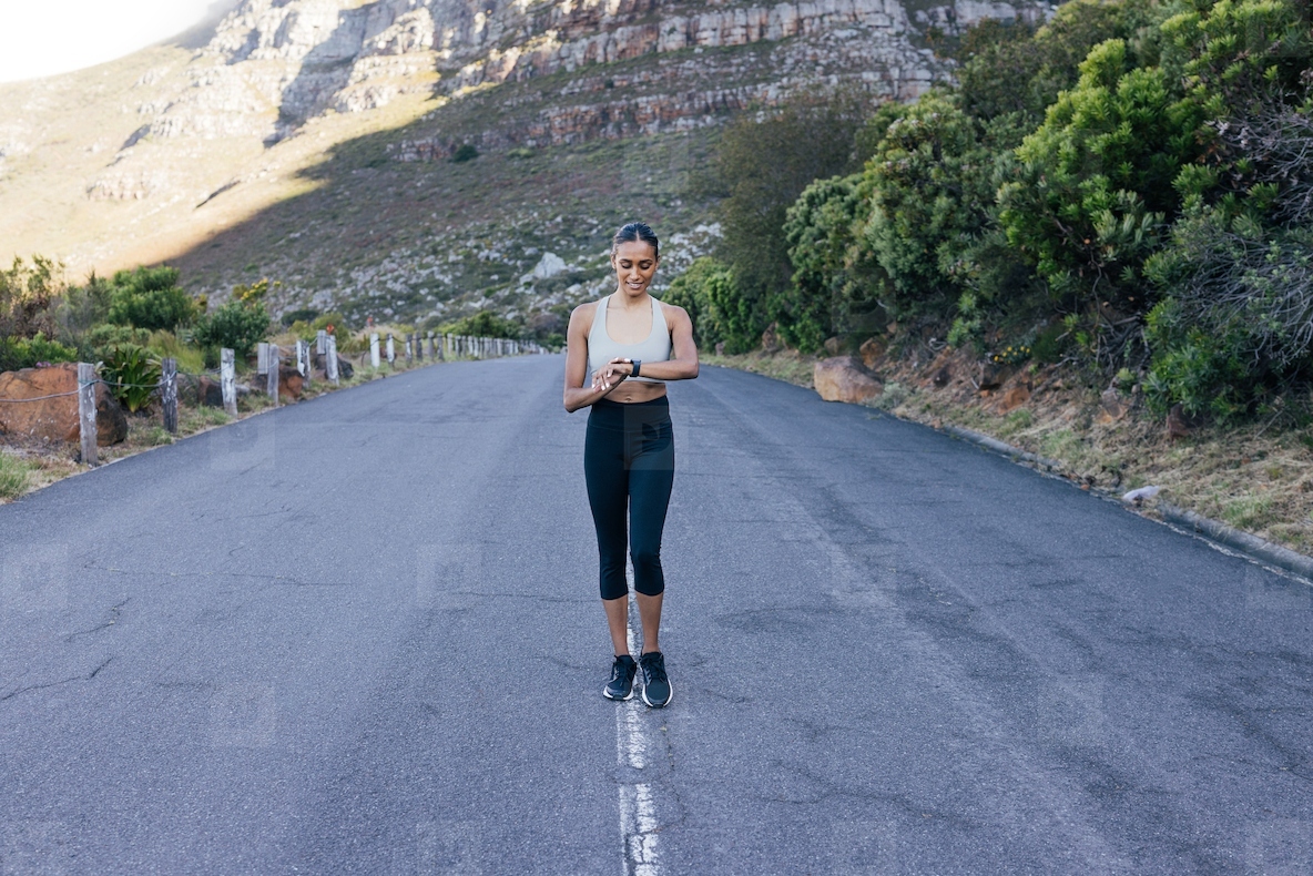 Full length of a smiling female walking on an abandoned road and checking her pulse Cheerful woman using smartwatches while doing morning walk