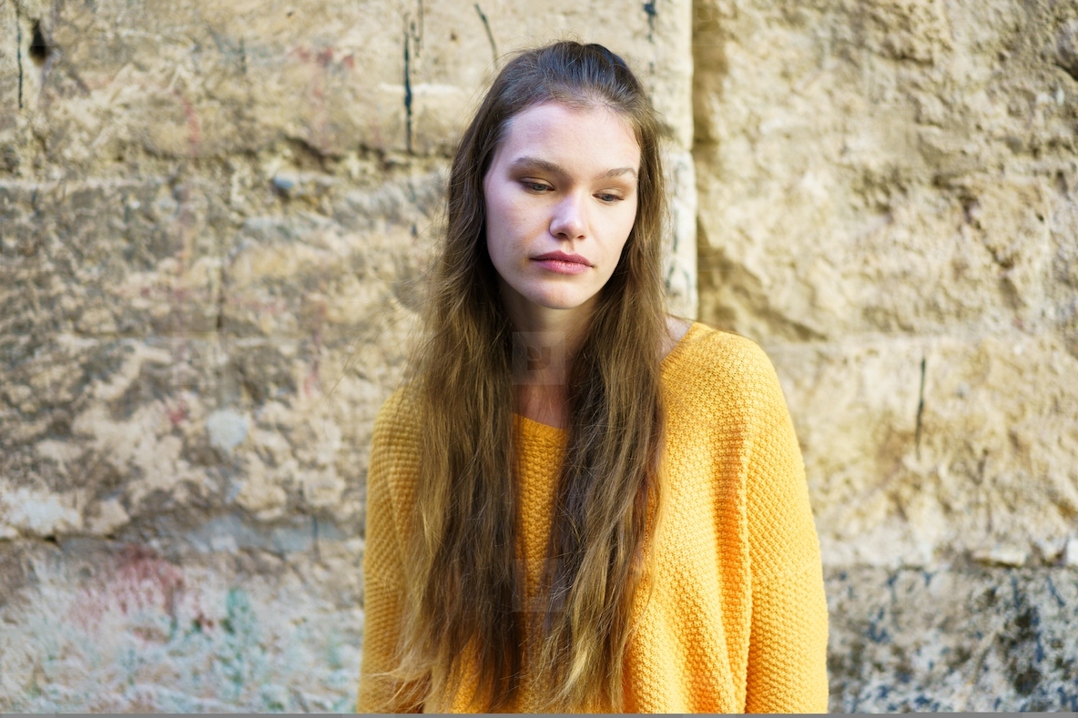 A contemplative woman in a bright yellow sweater poses gracefully by a rugged stone wall