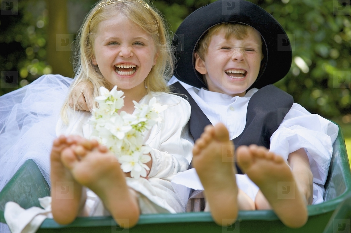 Boy and a girl wearing bride and groom costumes sitting in a wheelbarrow laughing