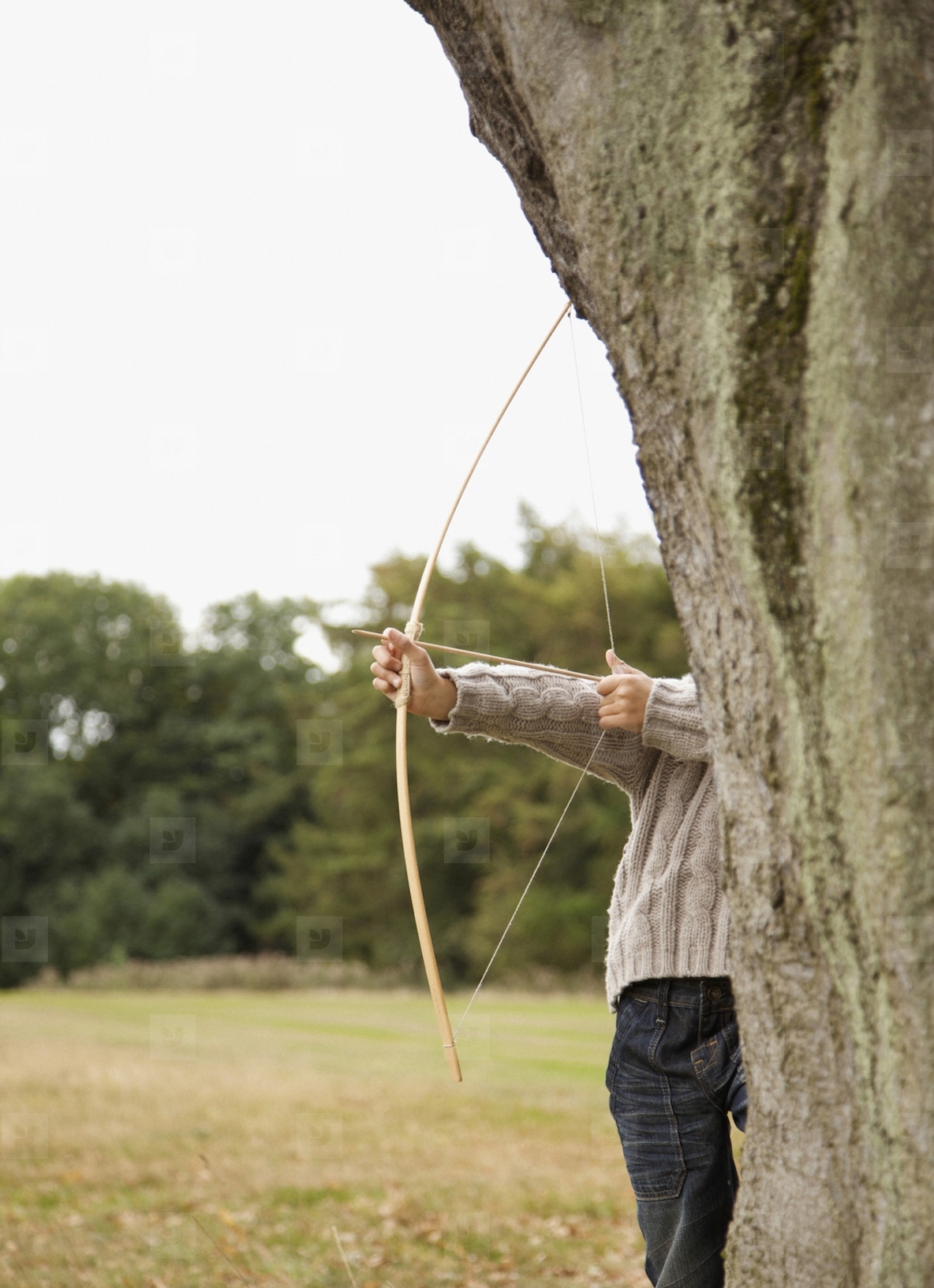 Young boy partially hidden by a tree aiming with a bow and arrow