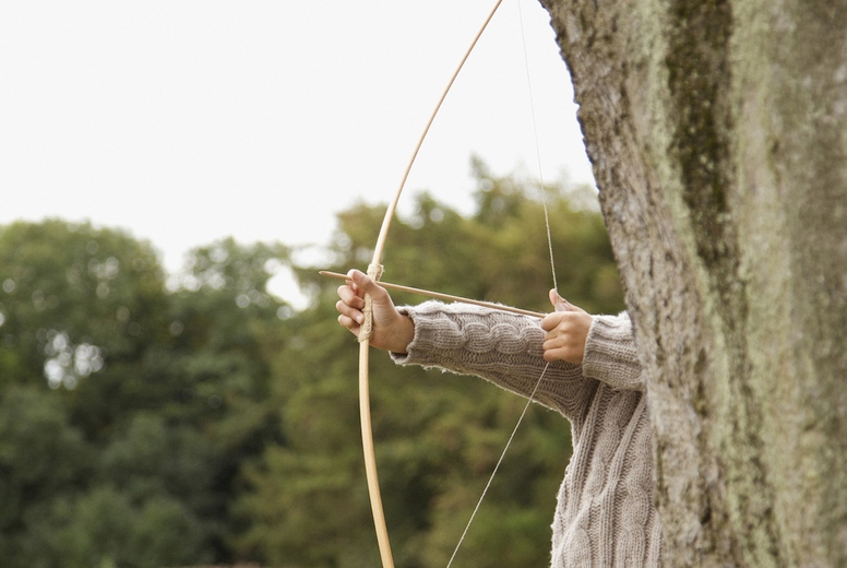 Young boy partially hidden by a tree aiming with a bow and arrow