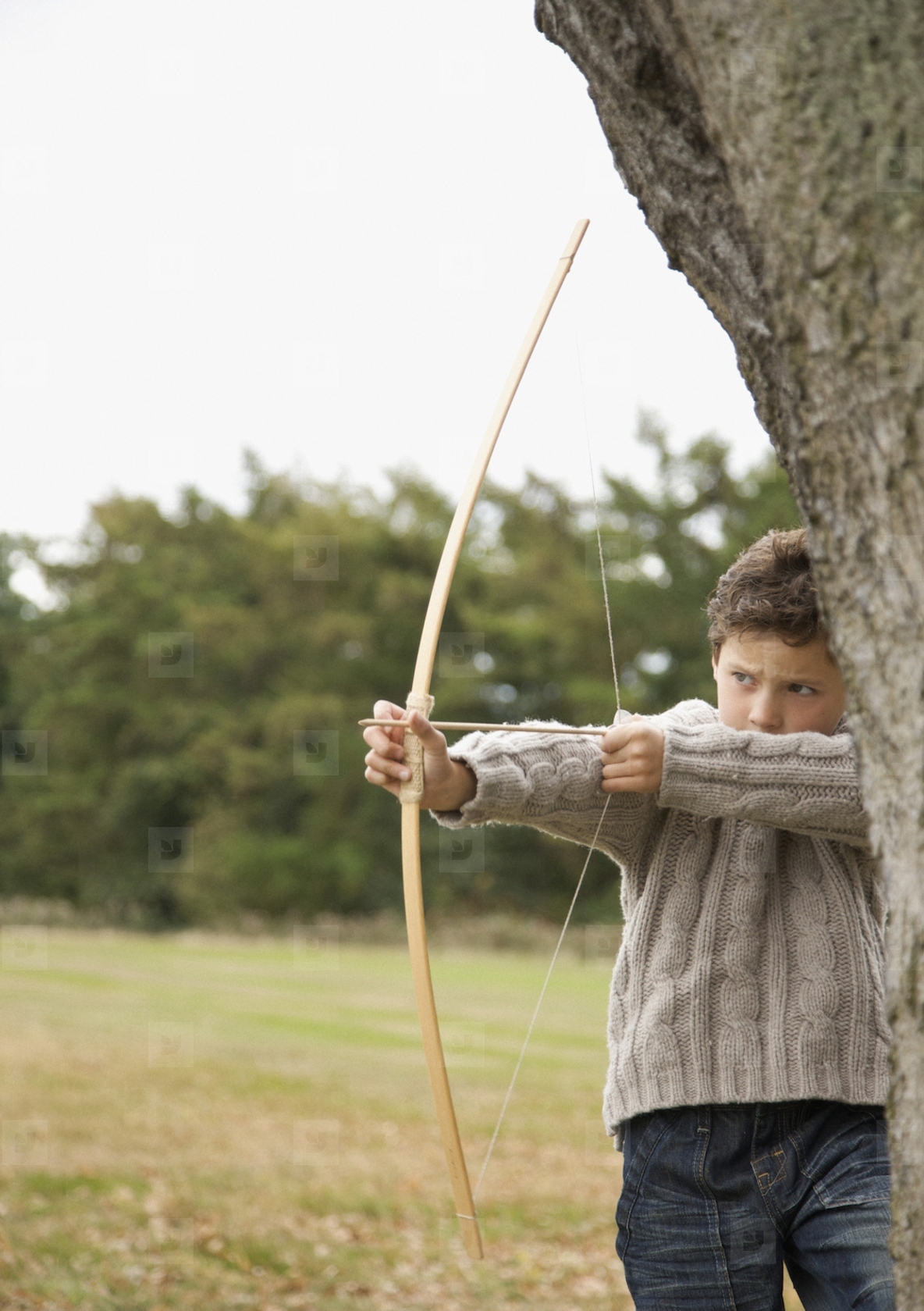 Young boy standing by a tree aiming with a bow and arrow