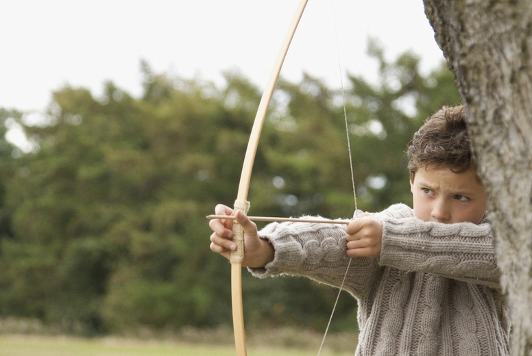 Young boy standing by a tree aiming with a bow and arrow
