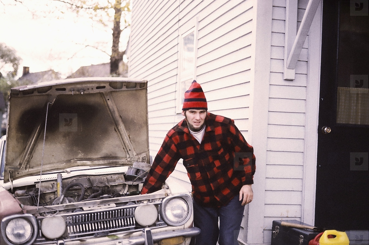 Portrait young man in stocking cap working on car engine in driveway