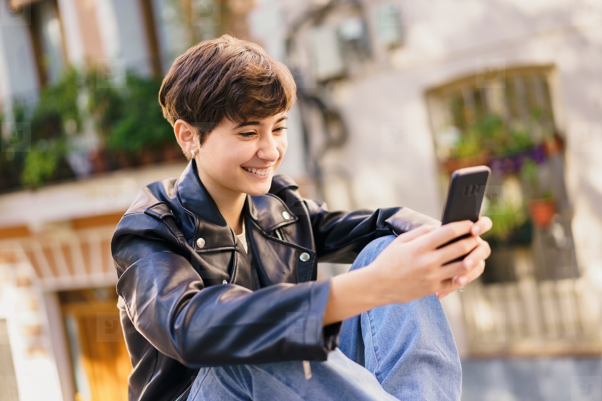 A cheerful young individual is actively engaged with their smartphone in a lively urban setting