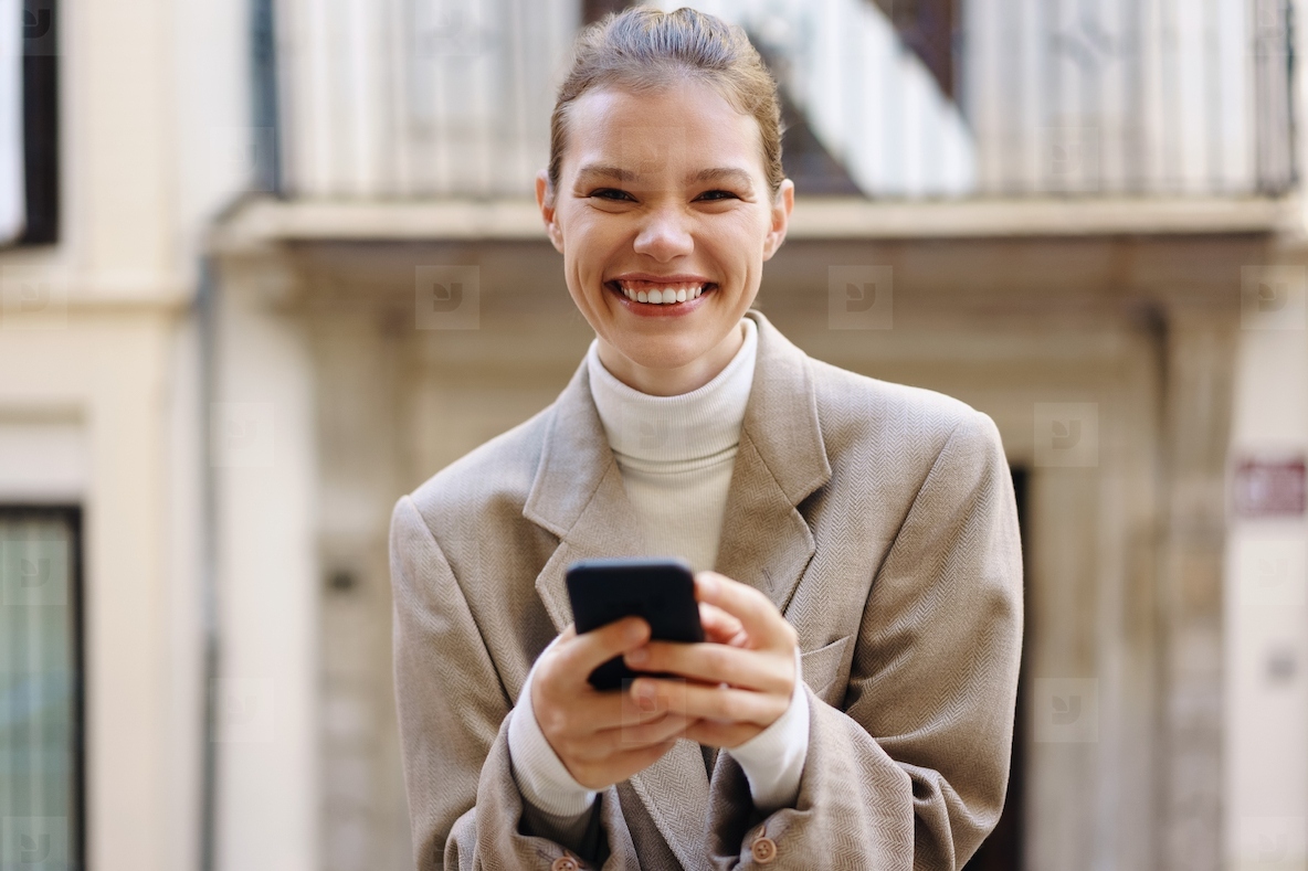 A joyful young woman is smiling brightly while she is texting on her smartphone outdoors