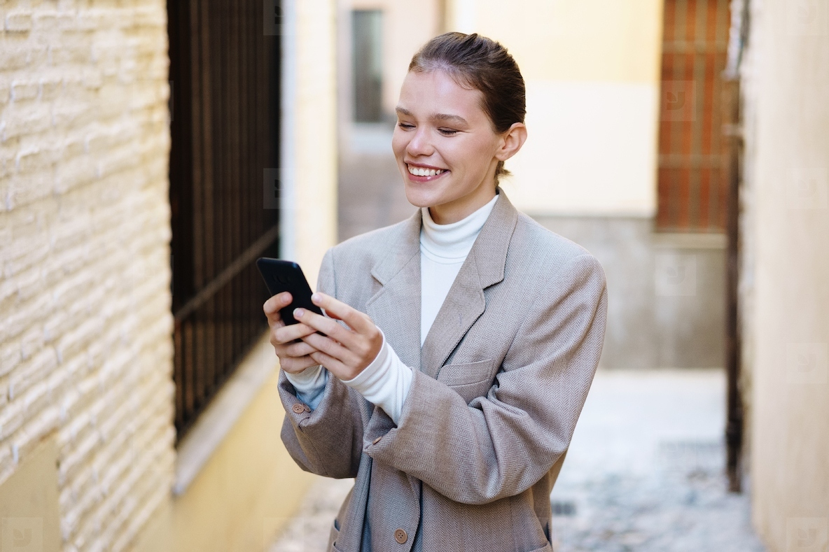 A cheerful young woman in a stylish outfit smiles while using her smartphone in a city alley