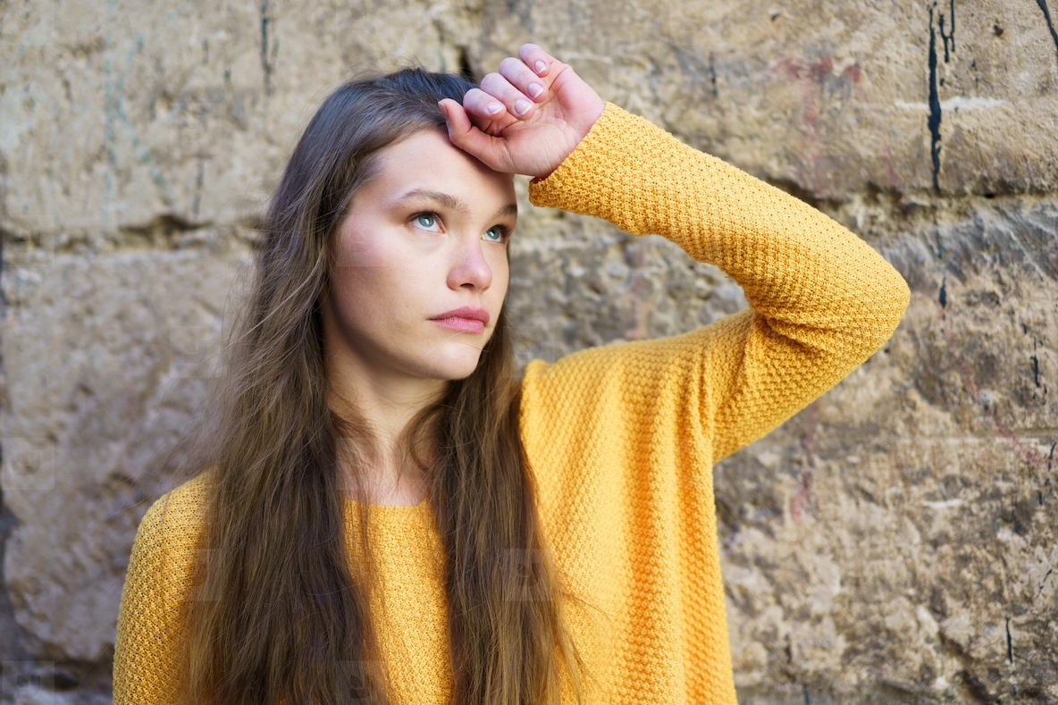 A young woman in a yellow sweater stands thoughtfully by a stone wall exuding calm elegance