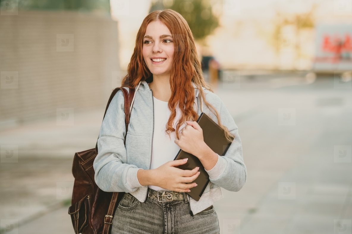 A smiling young woman joyfully carrying books while casually walking along an urban street