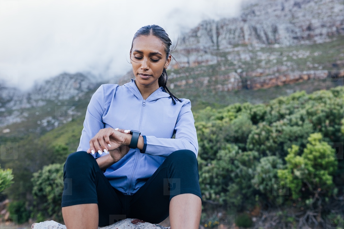 Female in sports clothes checking her smartwatches while sitting against a stunning view with mountains and clouds