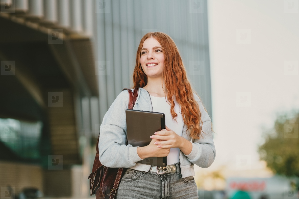 A young woman with a joyful smile is outdoors  holding a tablet and wearing a stylish backpack