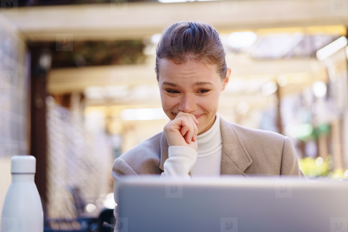 A young woman is smiling at her laptop in a modern outdoor setting enjoying her work