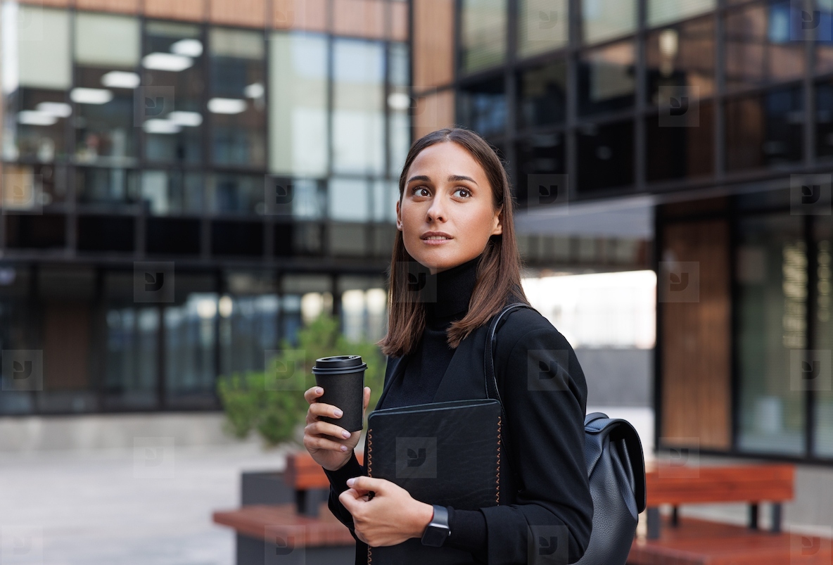 Young woman entrepreneur holding coffee to go wearing black clothes Stylish female is standing in the courtyard of an office building holding a digital tablet and looking away