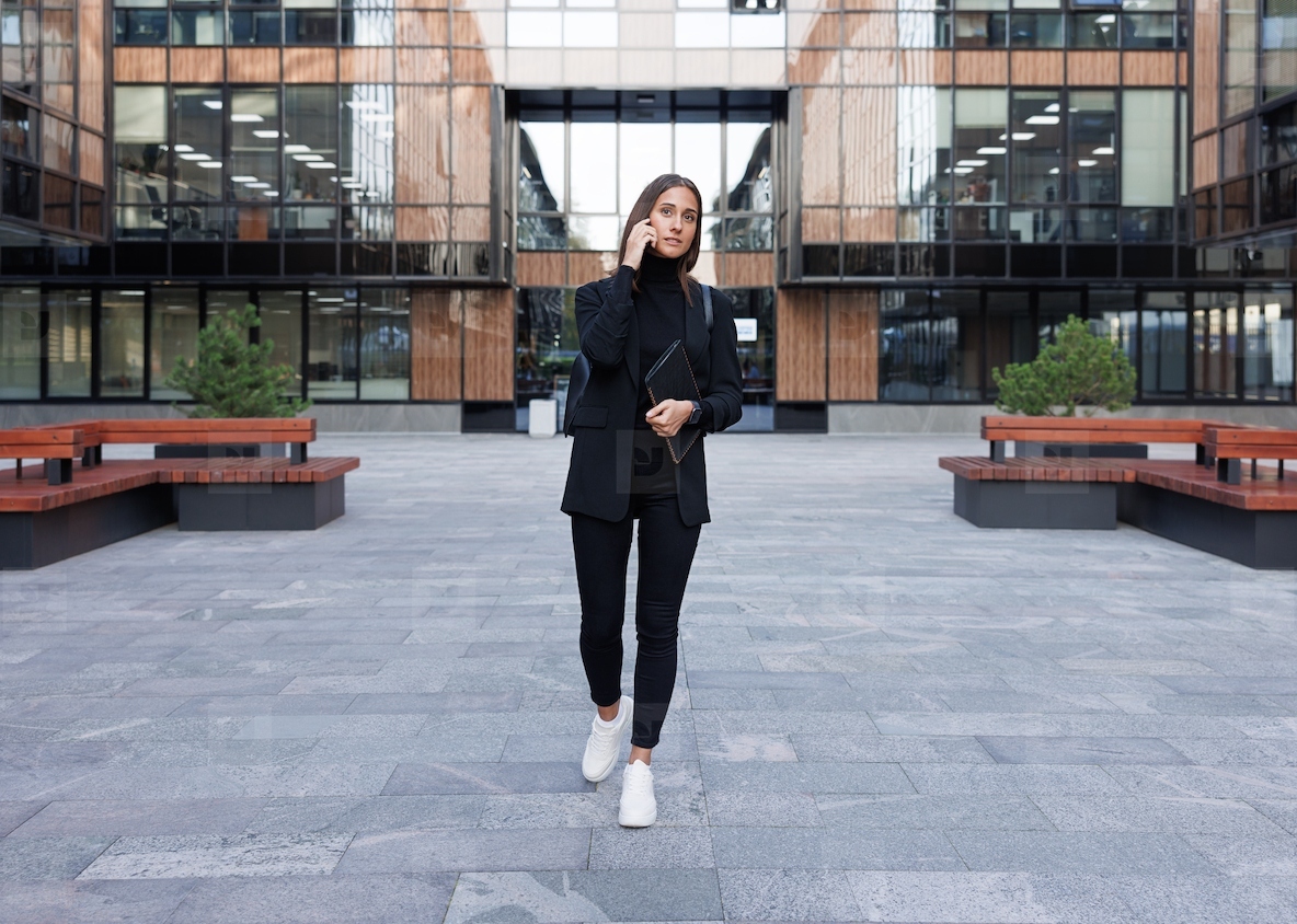 Full length of young woman in stylish black clothes walking in front of an office building  Female entrepreneur holding a digital tablet in case and talking on mobile phone while walking