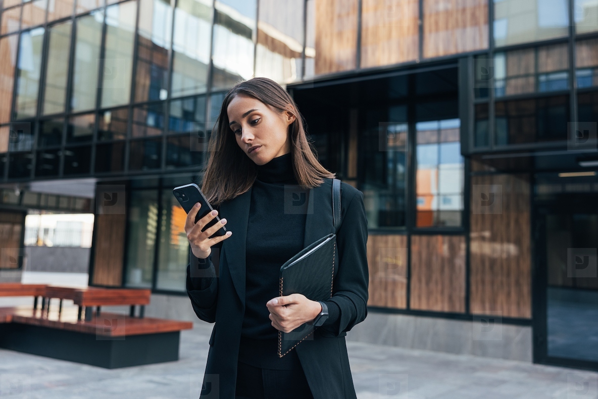 Confident and serious businesswoman looking at her smartphone while holding a digital tablet and standing against business building