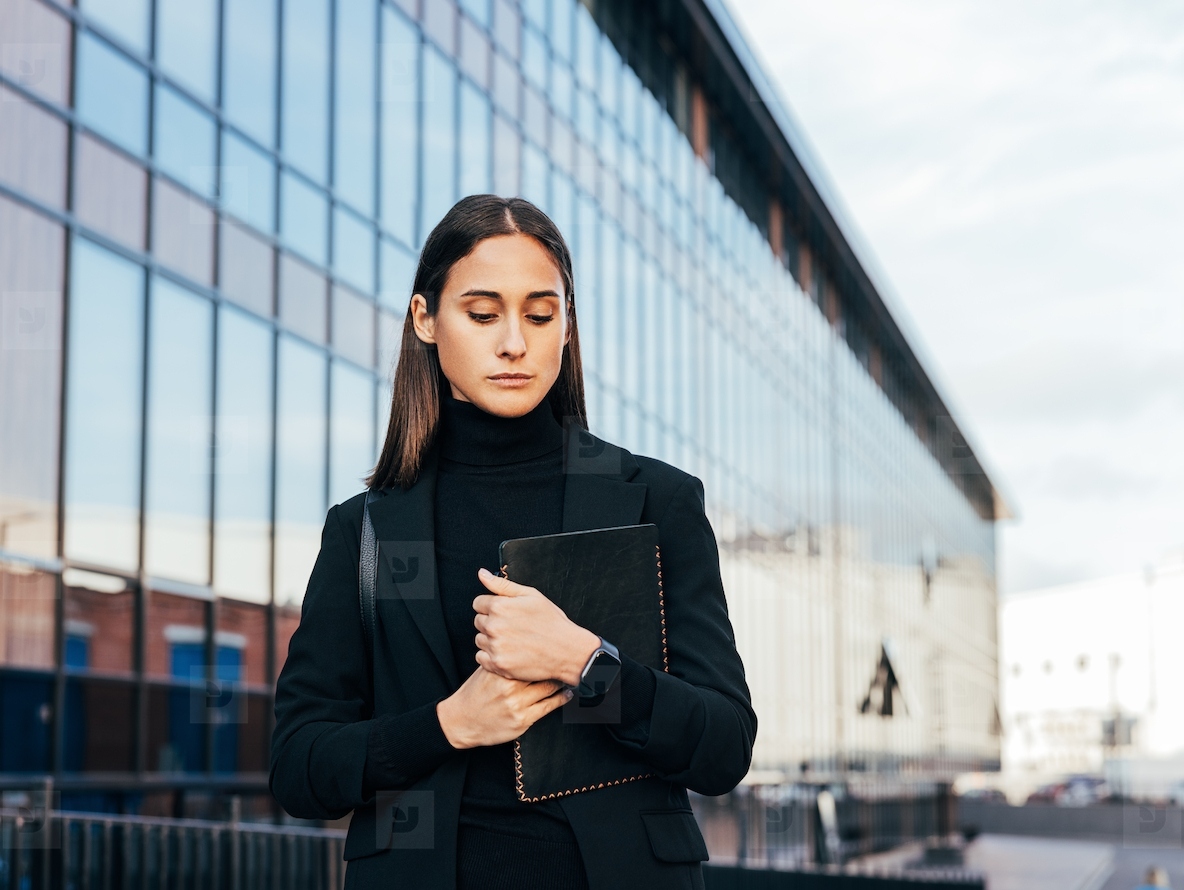 Confident businesswoman in black formal attire holding a digital tablet and looking down while standing outdoors