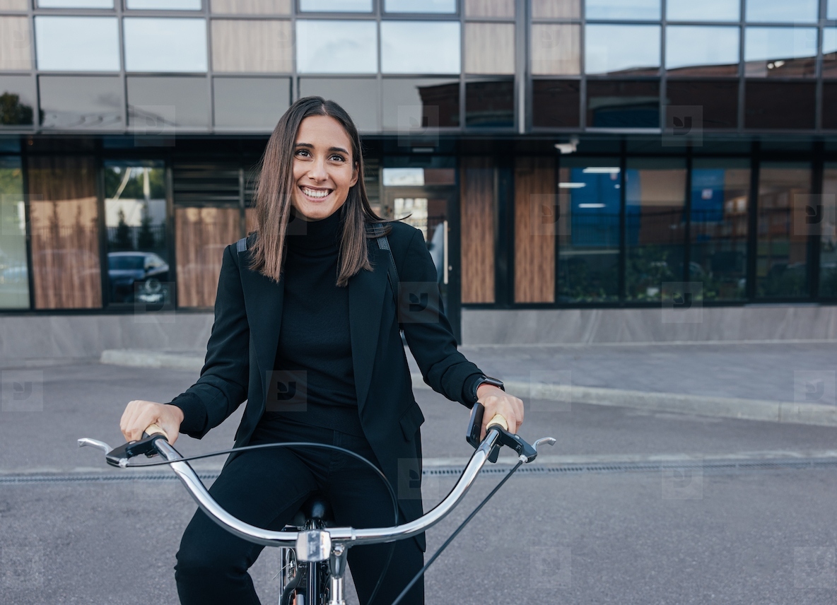 Young cheerful female in black clothes sitting on a bicycle outdoors  Stylish businesswoman in front of a glass building sitting on a bicycle