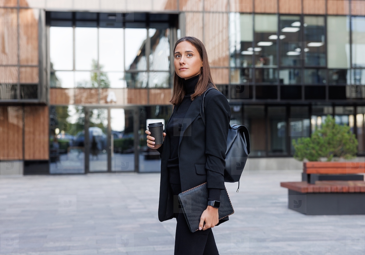 Side view of young female entrepreneur wearing black clothes with high collar Businesswoman with digital tablet and cup of coffee to go against glass building