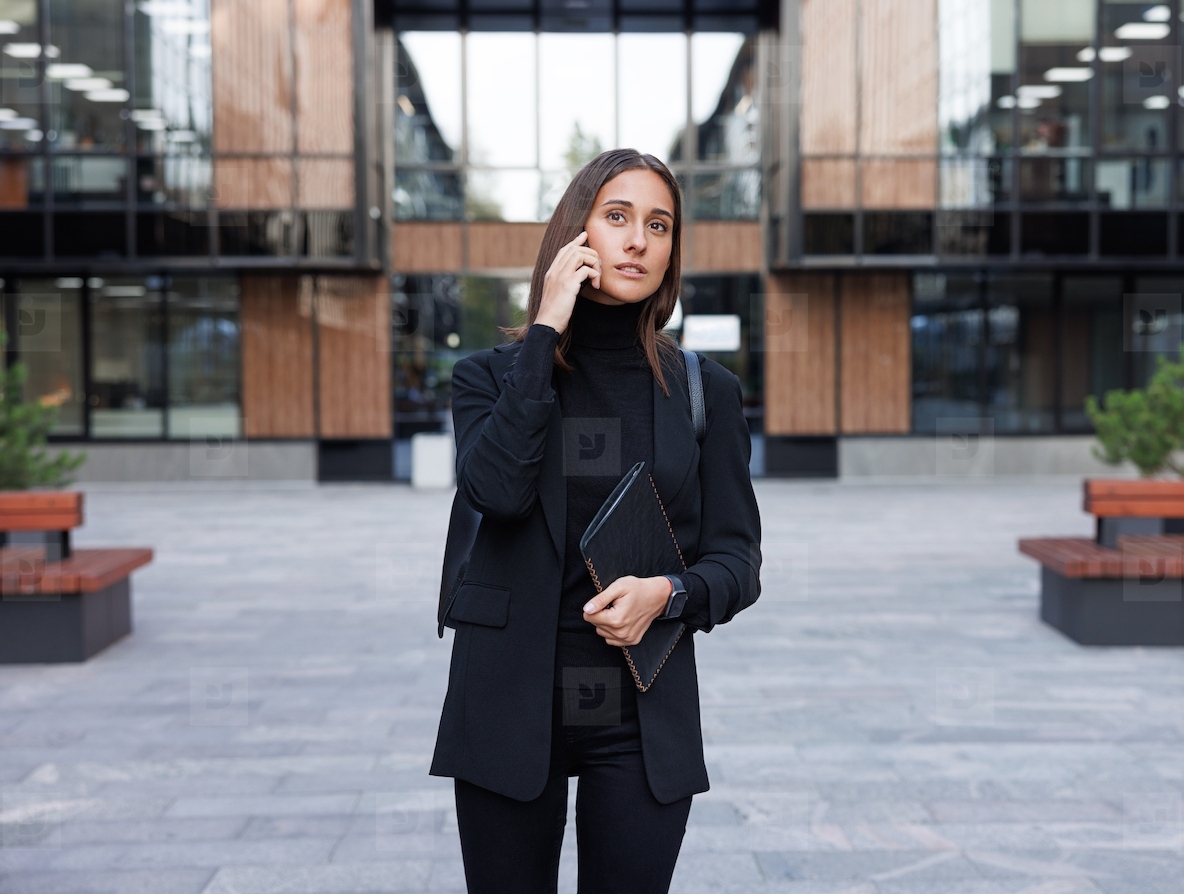 Young female entrepreneur wearing black formal stylish clothes with high collar making a phone call in country yard of an office building