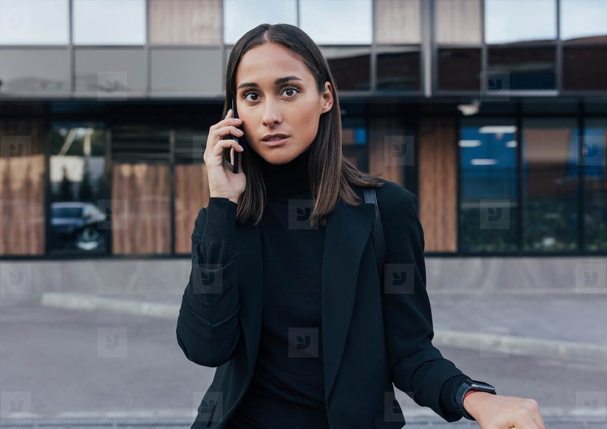 Portrait of a young female with smartphone wearing black formal attire and looking at camera while standing outside